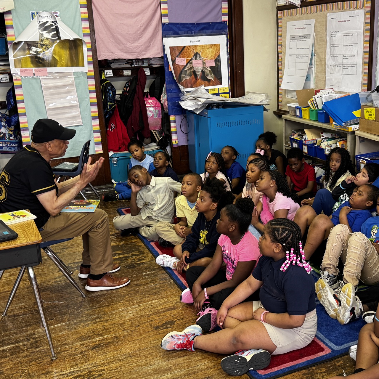 A guest speaker reads to a group of attentive students seated on the floor in a classroom at Overbrook Elementary School, encouraging curiosity and discussion during storytime.