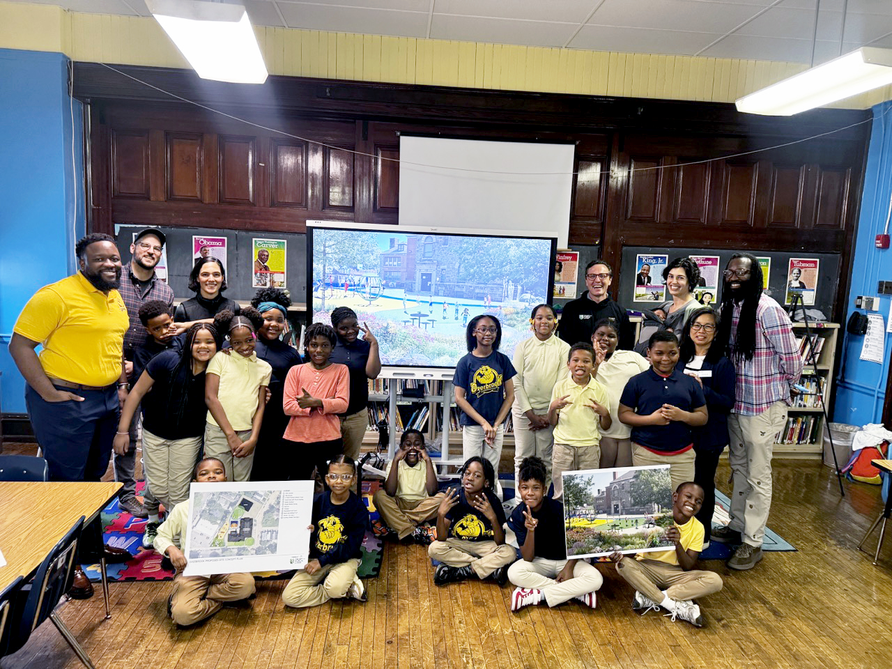 Students and educators gather in a classroom at Overbrook Elementary School to present design plans for a school improvement project, smiling together in front of a large screen and project posters.