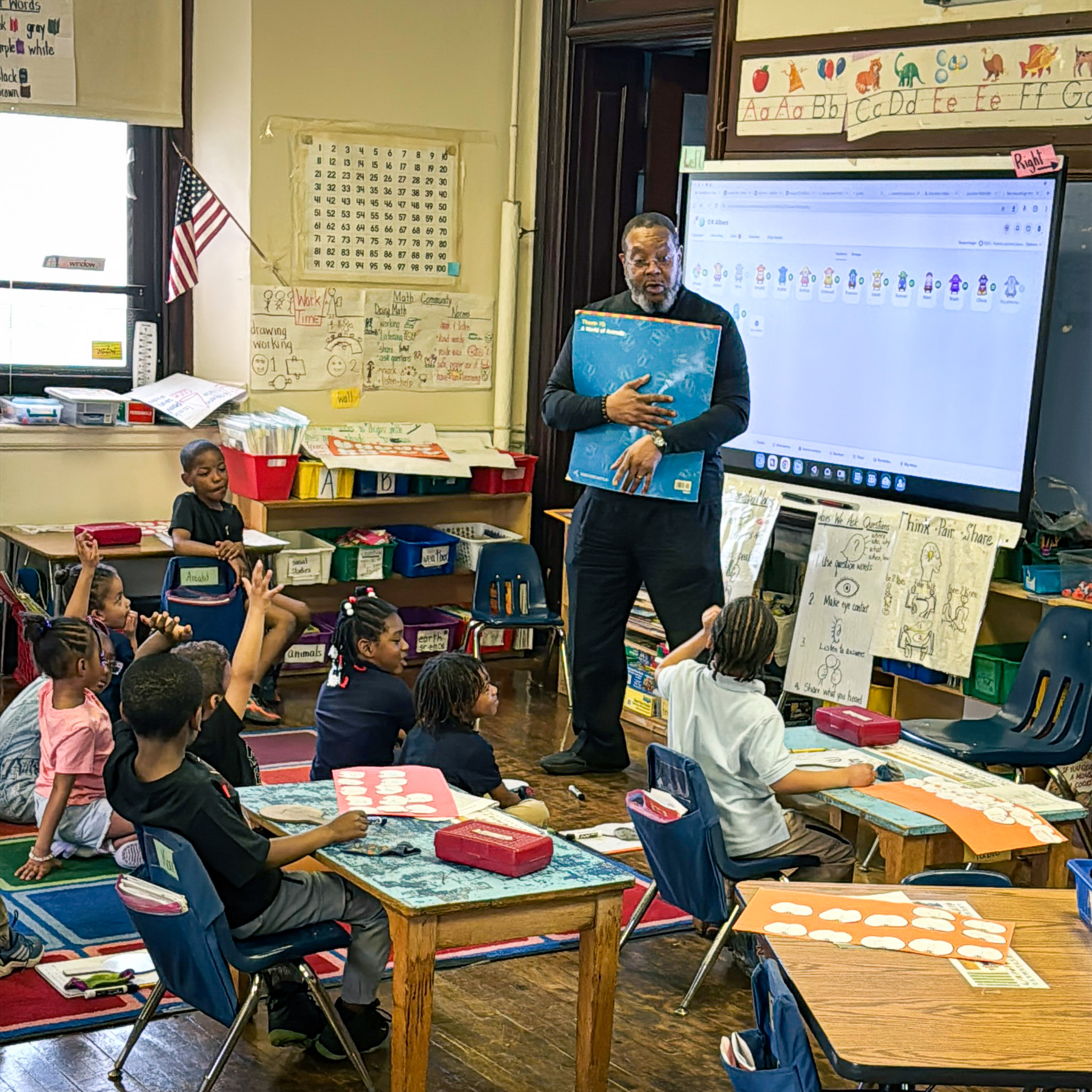 A teacher reads aloud to kindergarten students seated on the classroom rug at Overbrook Elementary School, engaging them in an interactive lesson surrounded by colorful charts and learning materials.